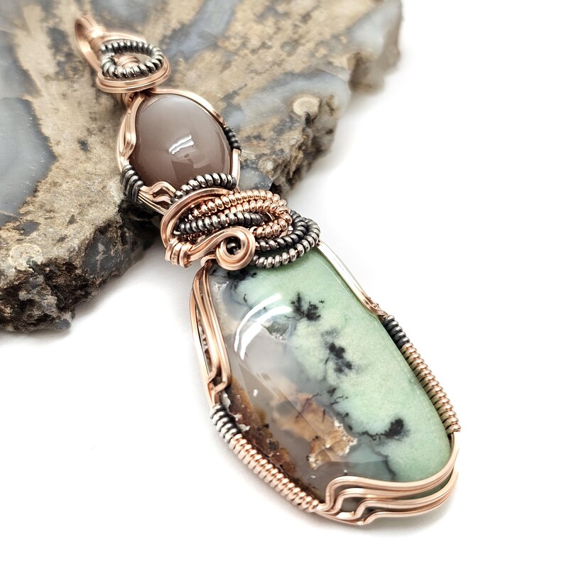 May include: A handcrafted pendant featuring a light green and brown stone, accented with black inclusions. The stone is encased in copper wire and a smaller, oval-shaped, gray stone is set above it. The pendant is displayed on a textured, gray surface.