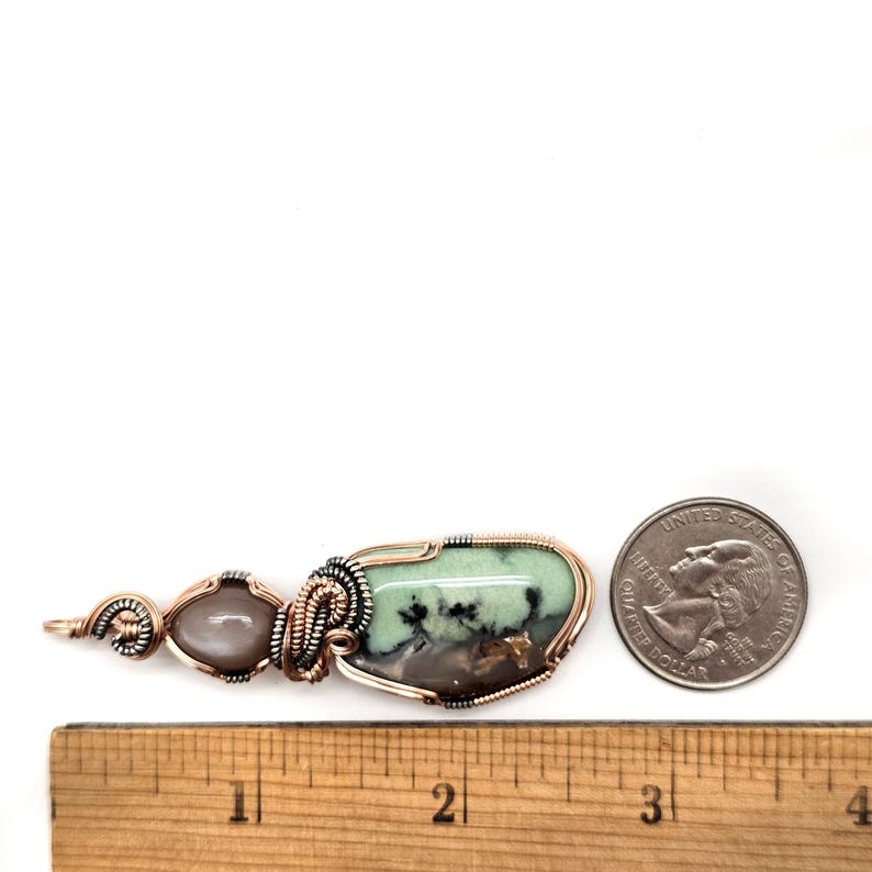 May include: A handcrafted pendant featuring a light green and brown stone, intricately wrapped in copper wire. A smaller, oval-shaped stone is also included. The pendant is next to a quarter and a ruler for size reference.