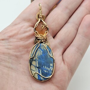 May include: A handcrafted pendant featuring a large, oval blue stone with white veining, accented by a smaller, orange-brown stone. Both are encased in gold-colored wire, creating a unique, artistic jewelry piece.