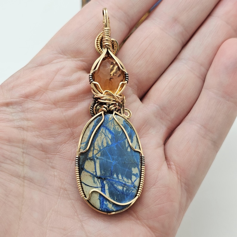 May include: A handcrafted pendant featuring a large, oval blue stone with white veining, accented by a smaller, orange-brown stone. Both are encased in gold-colored wire, creating a unique, artistic jewelry piece.