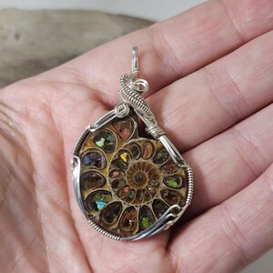 May include: A handcrafted pendant featuring a polished ammonite fossil encased in a silver wire wrap. The fossil displays a spiral pattern with iridescent colors of green, red, and gold. The pendant has a loop for a chain.