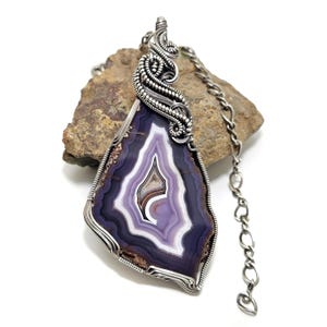 May include: A handcrafted pendant necklace featuring a large, polished agate stone with concentric bands of purple, white, and brown. The stone is encased in a silver wire wrap and hangs from a silver chain. The agate is approximately 3 inches long.