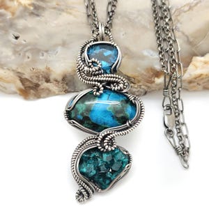 May include: A handcrafted silver pendant necklace featuring three gemstone pendants. The top pendant is a clear blue gemstone, the middle is a large oval blue and green stone, and the bottom is a cluster of teal crystals. The necklace chain is also silver.