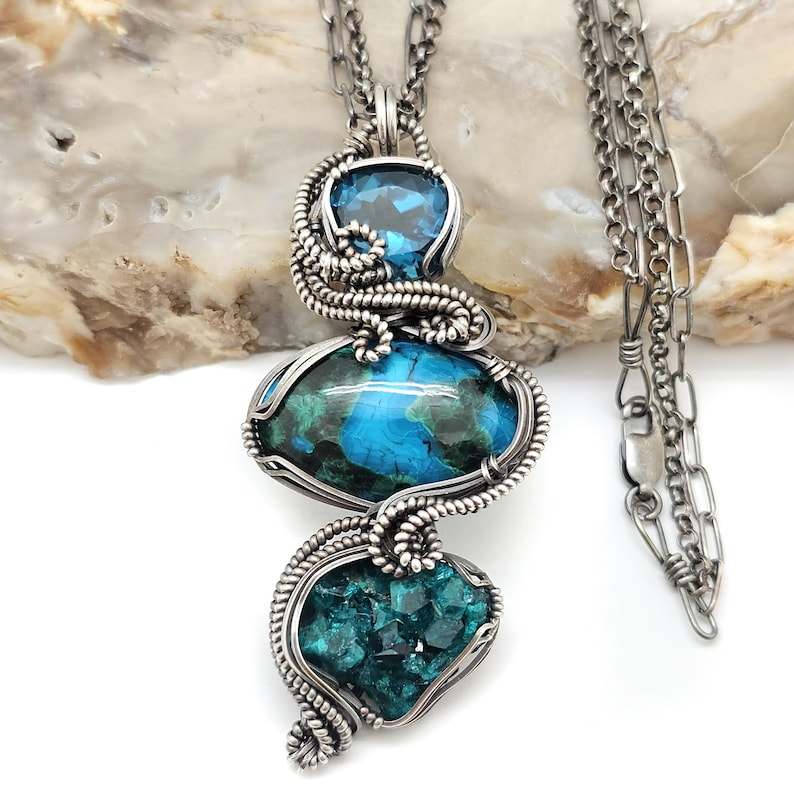 May include: A handcrafted silver pendant necklace featuring three gemstone pendants. The top pendant is a clear blue gemstone, the middle is a large oval blue and green stone, and the bottom is a cluster of teal crystals. The necklace chain is also silver.