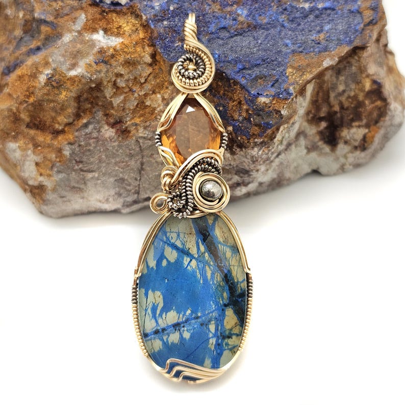 May include: A handcrafted pendant with a gold-colored wire wrap design. The pendant features a large, oval-shaped blue stone with a unique pattern, a faceted orange gemstone, and a small silver-colored bead. The top of the pendant has a spiral design.