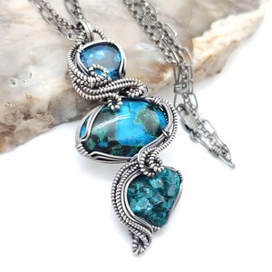 May include: A handcrafted silver pendant necklace featuring three vibrant gemstones. The necklace showcases a blue gemstone at the top, a larger oval blue-green stone in the center, and a teal stone at the bottom. The stones are intricately wire-wrapped.