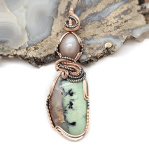 May include: A handcrafted pendant featuring a large, oval-shaped green and gray stone, accented with black inclusions. The stone is encased in copper wire with a silver-colored wire accent. A smaller, oval-shaped, gray stone is at the top.