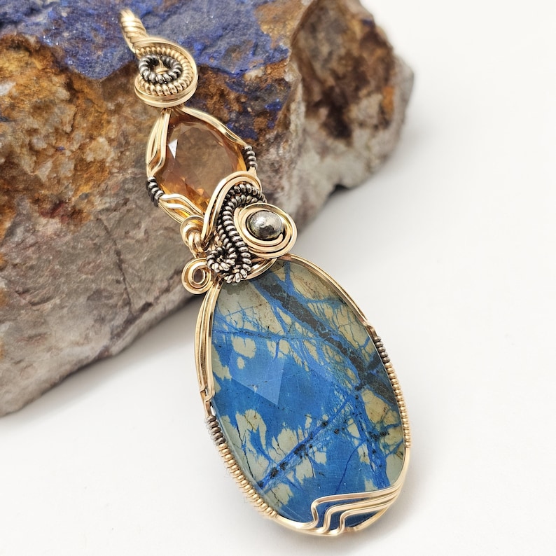 May include: A handcrafted pendant featuring a large, oval blue gemstone with a unique pattern, accented by a smaller amber-colored stone. The pendant is intricately wire-wrapped in gold and silver tones, creating a striking contrast.