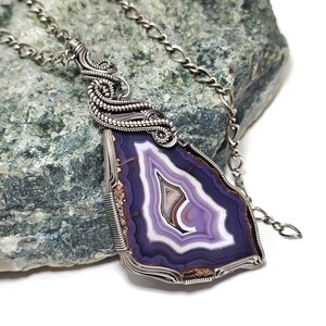 May include: A silver wire-wrapped pendant necklace featuring a polished, purple and white agate stone. The stone has concentric bands of color and is suspended from a delicate silver chain. The pendant is set against a textured, green-gray rock.