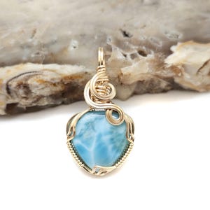 May include: A heart-shaped pendant featuring a light blue gemstone, intricately wrapped in gold-colored wire. The wirework forms a decorative bail and frame around the stone. The pendant is displayed against a neutral background.