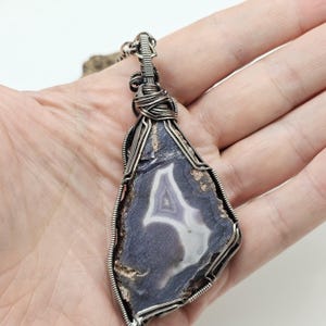May include: A handcrafted pendant necklace featuring a large, polished agate stone. The stone is a mix of deep blue, white, and brown hues, with a striking central pattern. The stone is encased in a silver-colored wire wrap, creating a unique and artistic piece.