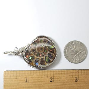 May include: A wire-wrapped pendant featuring a polished ammonite fossil. The fossil displays iridescent colors and intricate internal patterns. The pendant is next to a quarter coin and a ruler, showing the size of the pendant.