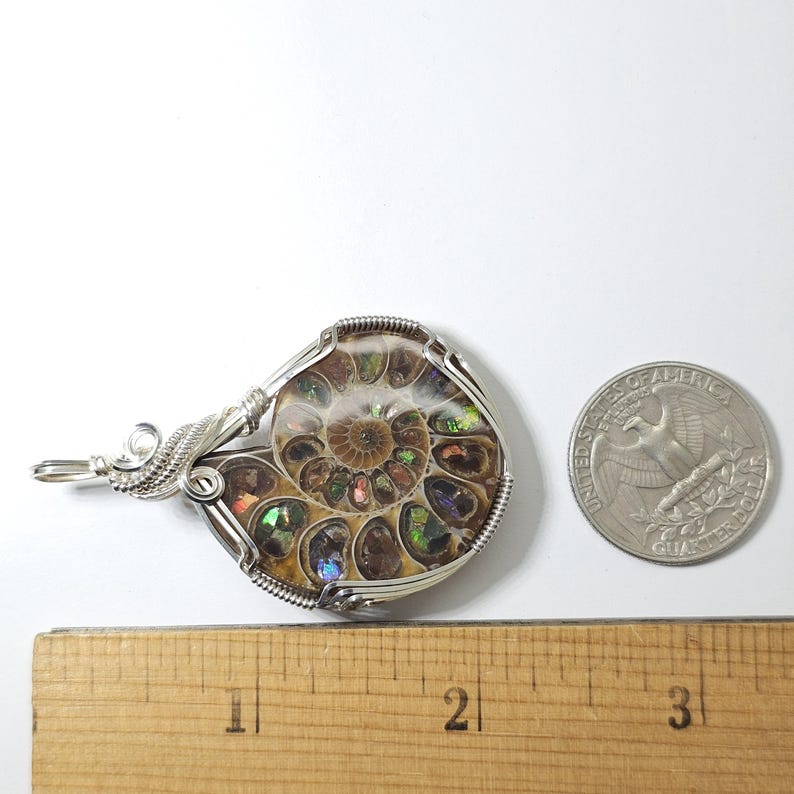 May include: A wire-wrapped pendant featuring a polished ammonite fossil. The fossil displays iridescent colors and intricate internal patterns. The pendant is next to a quarter coin and a ruler, showing the size of the pendant.