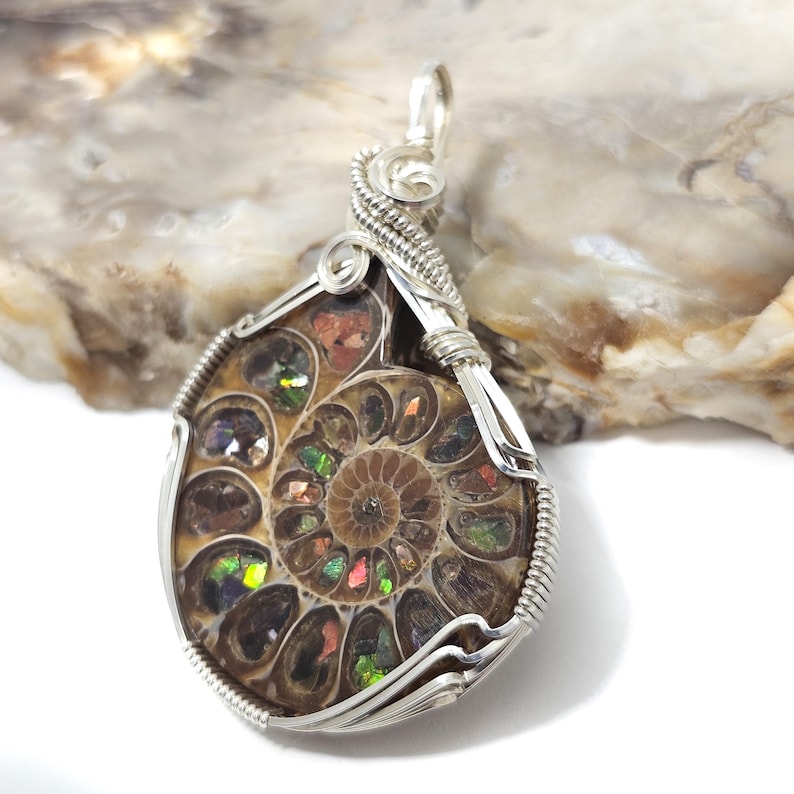 May include: A pendant featuring a polished ammonite fossil encased in a silver wire wrap. The fossil displays a spiral pattern with iridescent colors of green, red, and gold. The silver wire is intricately woven around the fossil, creating a unique jewelry piece.