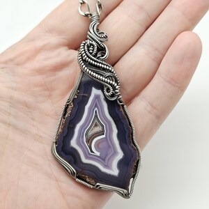 May include: A handcrafted pendant featuring a polished, teardrop-shaped agate stone with concentric bands of purple, white, and brown. The stone is encased in a silver wire wrap with intricate detailing, including a decorative swirl at the top and a bail for a necklace chain.