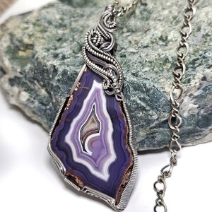 May include: A handcrafted pendant necklace featuring a polished, teardrop-shaped agate stone with layers of purple, white, and brown hues. The stone is encased in a silver wire wrap and hangs from a silver chain. The agate measures approximately 2 inches in length.