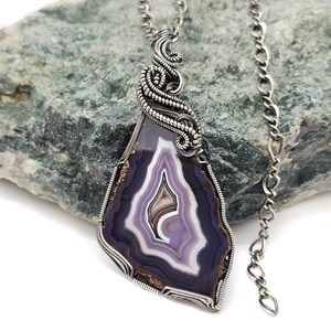 May include: A pendant necklace featuring a polished agate stone with a striking pattern of purple, white, and brown bands. The stone is encased in a silver wire wrap and hangs from a delicate silver chain. The pendant is displayed against a textured green stone.