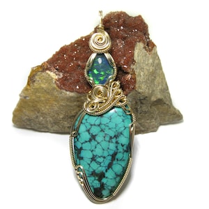 May include: A turquoise and green gemstone pendant with gold wire wrapping. The pendant is shaped like a teardrop and is set on a brown rock.