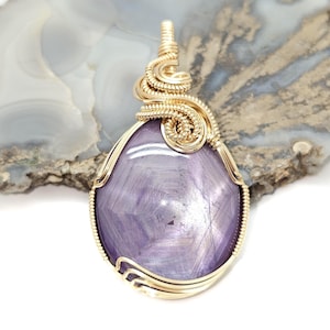May include: A gold pendant with a large, oval, lavender gemstone. The gemstone has a star pattern. The pendant is wire-wrapped in a decorative design. The pendant is displayed on a light-colored surface.