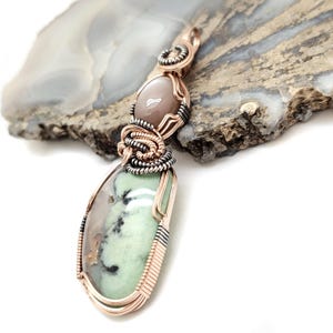 May include: A handcrafted pendant featuring two gemstones. The top stone is oval-shaped and light brown, while the bottom stone is a teardrop shape with green and gray hues. The stones are intricately wrapped with copper and silver wire.