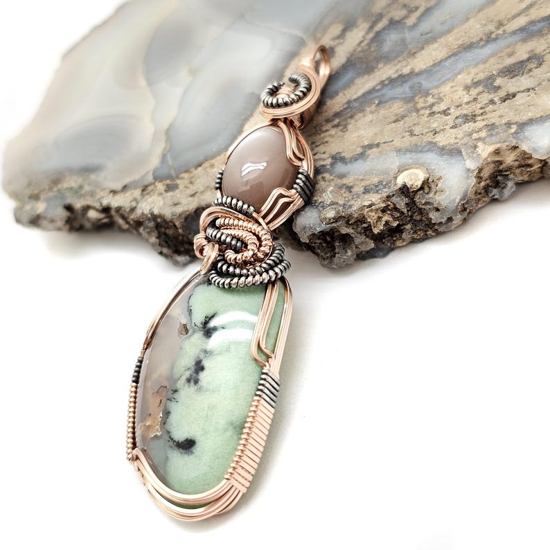 May include: A handcrafted pendant featuring two gemstones. The top stone is oval-shaped and light brown, while the bottom stone is a teardrop shape with green and gray hues. The stones are intricately wrapped with copper and silver wire.
