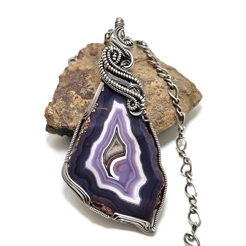 May include: A handcrafted pendant necklace featuring a polished, purple and white agate stone. The stone is encased in a silver wire wrap with intricate detailing. The necklace includes a silver chain, showcasing a unique, natural gemstone pendant.