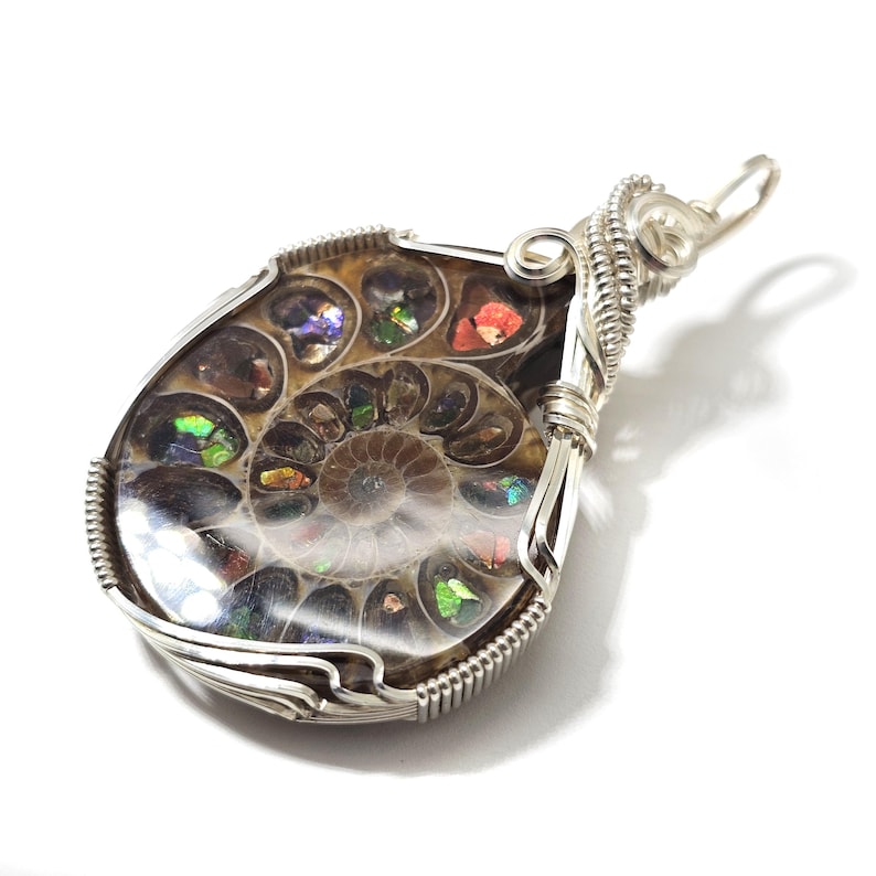 May include: A teardrop-shaped pendant featuring an iridescent ammonite fossil encased in a silver wire wrap. The fossil displays a spiral pattern with colorful, shimmering chambers. The pendant has a silver bail for hanging.