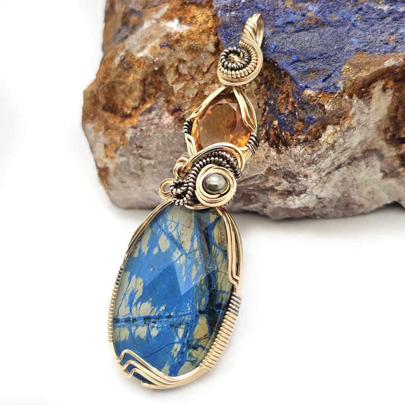 May include: A handcrafted pendant featuring a large, blue and gold stone, accented by a smaller amber-colored stone. The stones are encased in a gold-colored wire setting with intricate detailing, creating a unique jewelry piece.