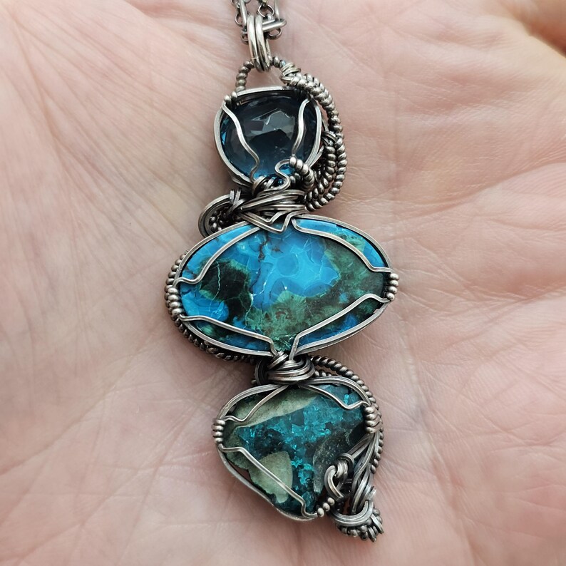 May include: A handcrafted pendant necklace featuring three gemstone elements. The top stone is a faceted blue crystal, followed by an oval-shaped stone with blue and green patterns, and a heart-shaped stone with similar colors. The stones are wrapped in silver wire.