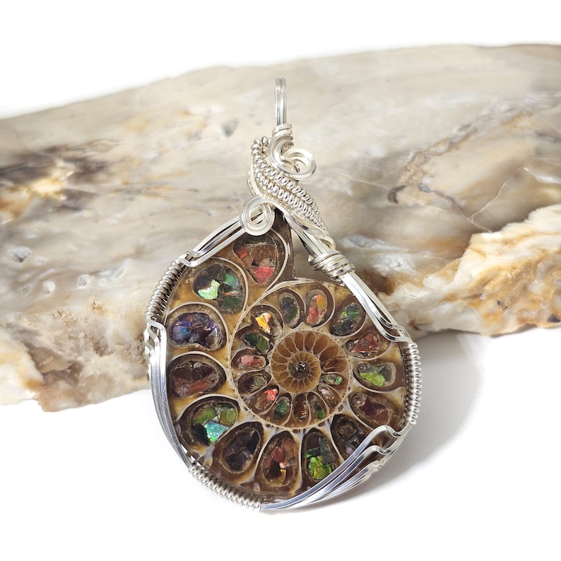 May include: A wire-wrapped pendant featuring a polished ammonite fossil. The fossil displays a spiral pattern with iridescent colors of red, green, and gold. The pendant is encased in silver-colored wire, with a loop for a chain.