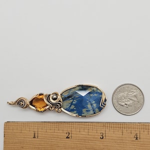 May include: A handcrafted pendant featuring a large, faceted blue gemstone with gold wire wrapping. An orange-yellow gemstone and a silver bead are also incorporated into the design. The pendant is next to a quarter for size comparison.