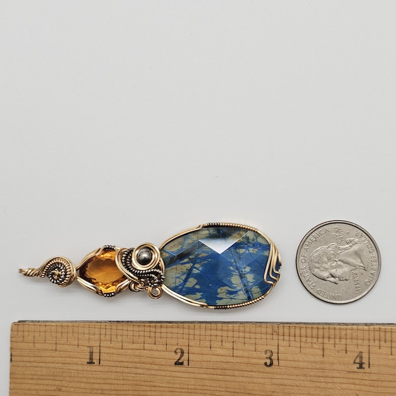 May include: A handcrafted pendant featuring a large, faceted blue gemstone with gold wire wrapping. An orange-yellow gemstone and a silver bead are also incorporated into the design. The pendant is next to a quarter for size comparison.