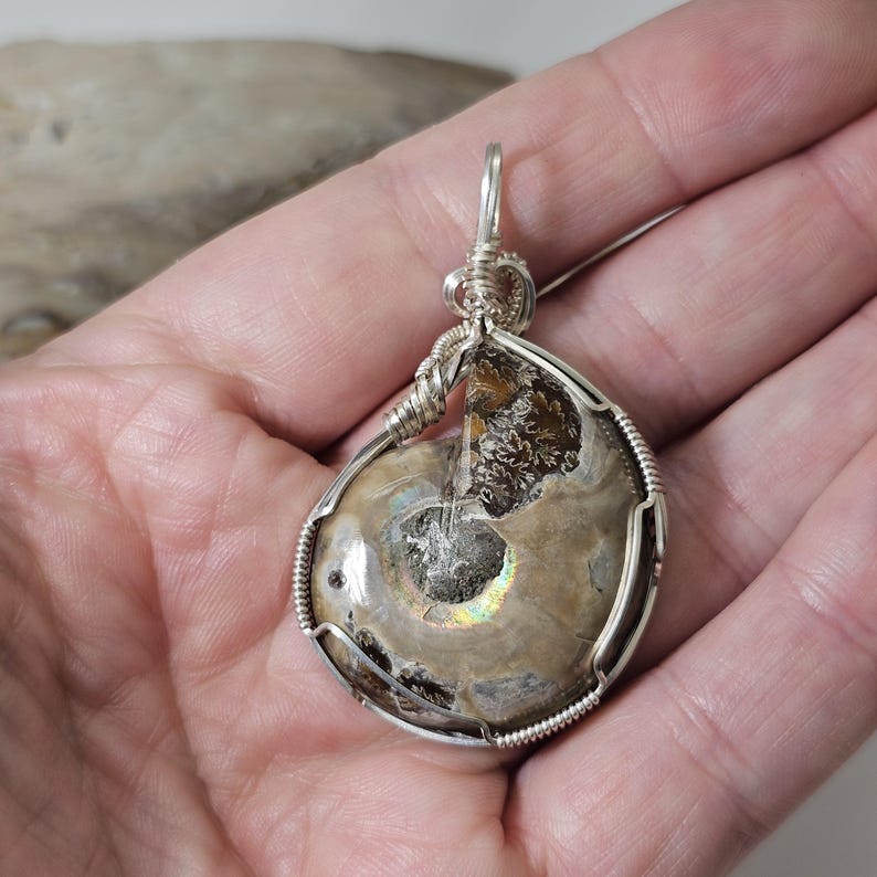 May include: A handcrafted pendant featuring a polished ammonite fossil encased in a silver wire wrap. The fossil displays a spiral pattern with iridescent highlights. The pendant is suspended from a silver bail.