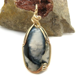 May include: A gold wire wrapped pendant featuring a gray and white agate gemstone. The pendant has a teardrop shape and is set in a delicate gold wire frame.