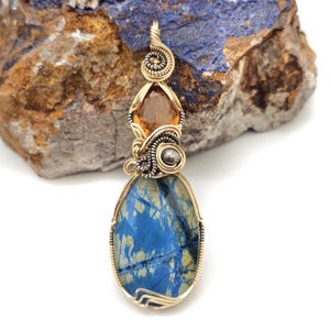 May include: A handcrafted pendant featuring a large, oval blue stone with a unique pattern, accented by a smaller orange gemstone and a silver-colored bead. The pendant is intricately wire-wrapped in gold-colored metal, showcasing a spiral design at the top.