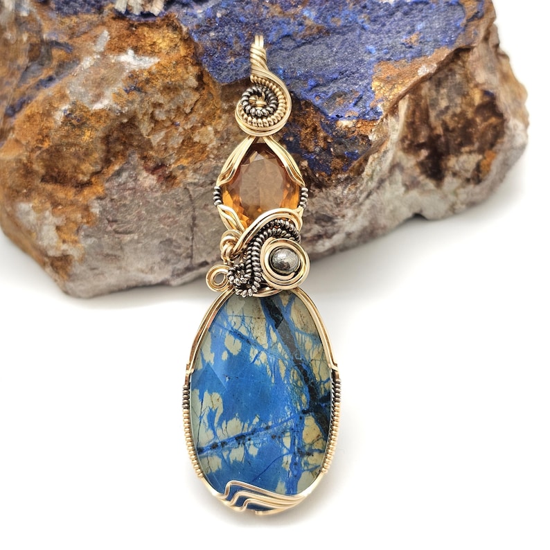 May include: A handcrafted pendant featuring a large, oval blue stone with a unique pattern, accented by a smaller orange gemstone and a silver-colored bead. The pendant is intricately wire-wrapped in gold-colored metal, showcasing a spiral design at the top.