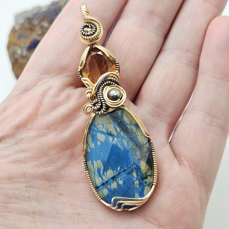 May include: A handcrafted pendant featuring a large, faceted blue stone with gold wire wrapping. The pendant includes a smaller amber-colored stone and a silver-colored accent. The wirework has a spiral design at the top.