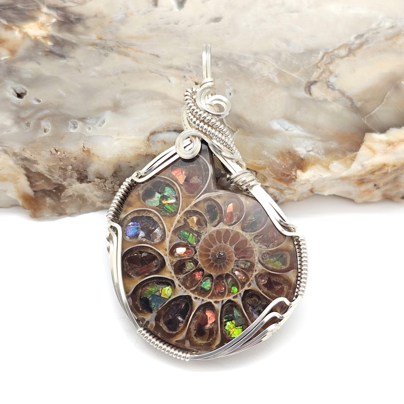 May include: A pendant featuring a polished ammonite fossil encased in a silver wire wrap. The fossil displays a spiral pattern with iridescent colors of green, blue, and red. The pendant has a loop for a chain.