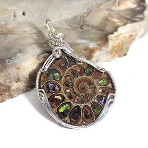 May include: A pendant featuring an iridescent ammonite fossil encased in a silver wire wrap. The fossil displays a spiral pattern with various colors, including green, blue, and brown. The silver wire is intricately woven around the fossil, creating a decorative frame.