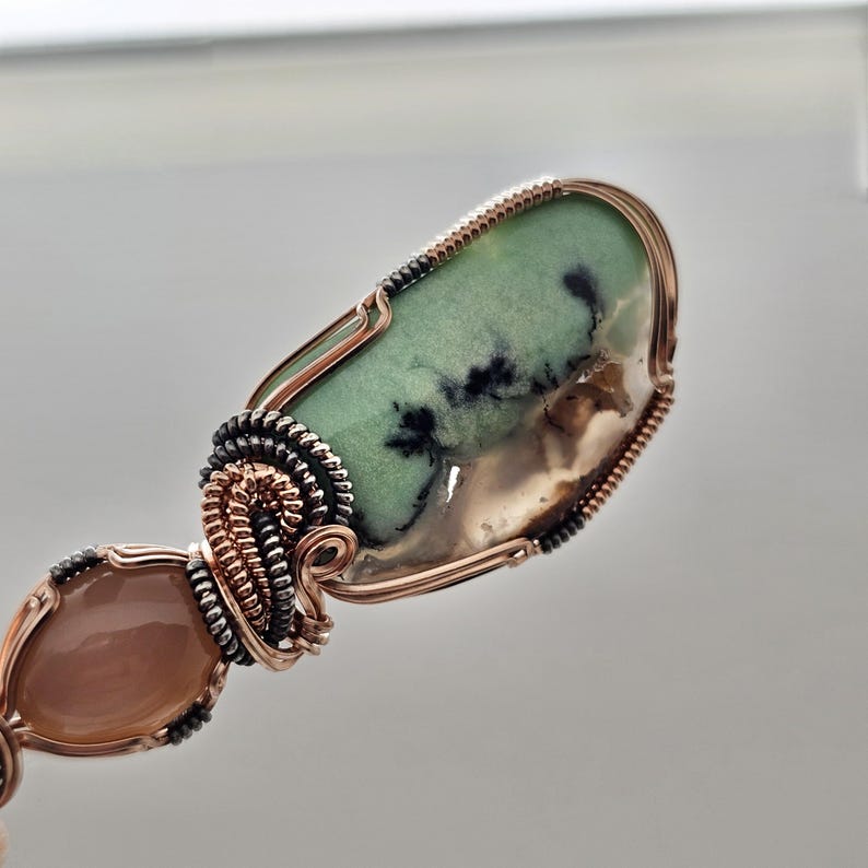 May include: A handcrafted pendant featuring a large, oval-shaped green stone with black inclusions, wire-wrapped in copper. Below, a smaller, teardrop-shaped peach-colored stone is also wire-wrapped. The intricate wirework adds a unique, artistic touch.