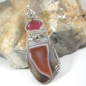 May include: A silver wire wrapped pendant featuring a red and white agate stone with a smaller pink stone at the top.