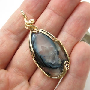 May include: Gold wire wrapped pendant featuring a gray and white agate gemstone with a unique pattern.