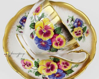 May include: A white teacup and saucer set with a gold rim and handle. The set is decorated with yellow, purple, and blue pansies. The saucer has a scalloped edge. A beautiful floral tea set.