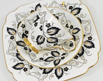May include: A white teacup, saucer, and square plate set with a black and gold leaf design. The teacup has a gold handle and rim. The set is on a white surface.
