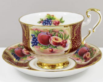 May include: A vintage teacup and saucer set. The cup and saucer are white with a gold trim and a detailed fruit design featuring peaches, grapes, and cherries. The cup has a decorative handle.