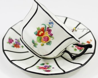 May include: A white teacup and saucer with black trim and floral designs. The cup has a black handle and the saucer has a scalloped edge.