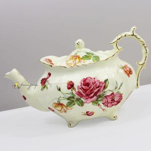 May include: Cream-colored porcelain teapot with a floral rose design. The teapot features a curved handle, a spout, and a lid with a small knob. The roses are in shades of pink, red, and yellow, with green leaves. Gold trim accents the edges.
