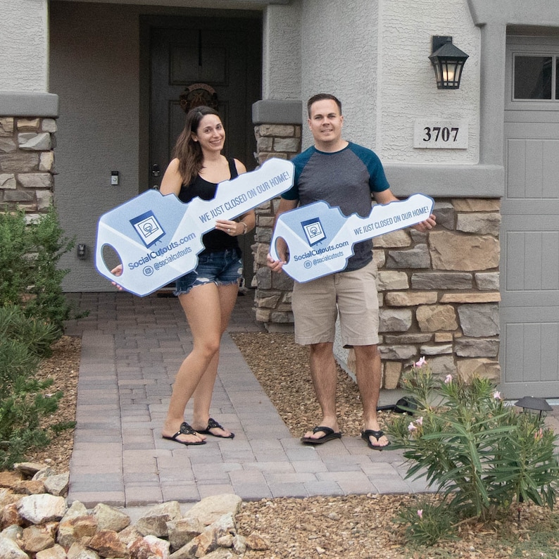 The photo is a man and woman holding signs. The signs say "WE JUST CLOSED ON OUR HOME!" and "SocialCutouts com @socialcutouts".