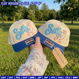 Op de afbeelding: Twee baseballpetten met een crèmekleurige kroon en een blauwe klep. Het woord "Silly" is lichtblauw geborduurd, met een roze rolschaatsende kippengrafiek. De tekst "FLAT VERSION - 3 SIZE 4" 5" 6"" staat bovenaan.