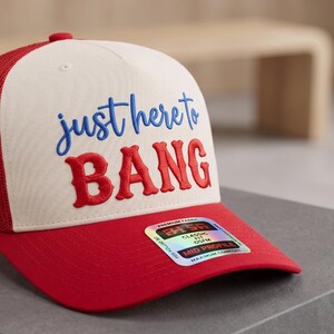 May include: A red and white trucker hat with the text "just here to BANG" embroidered in blue and red. The hat has a red brim and mesh back, and a small red button on top.